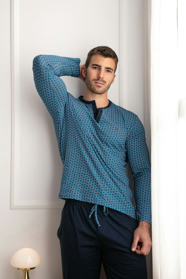 Man wearing a blue patterned shirt and dark pants against a white wall.